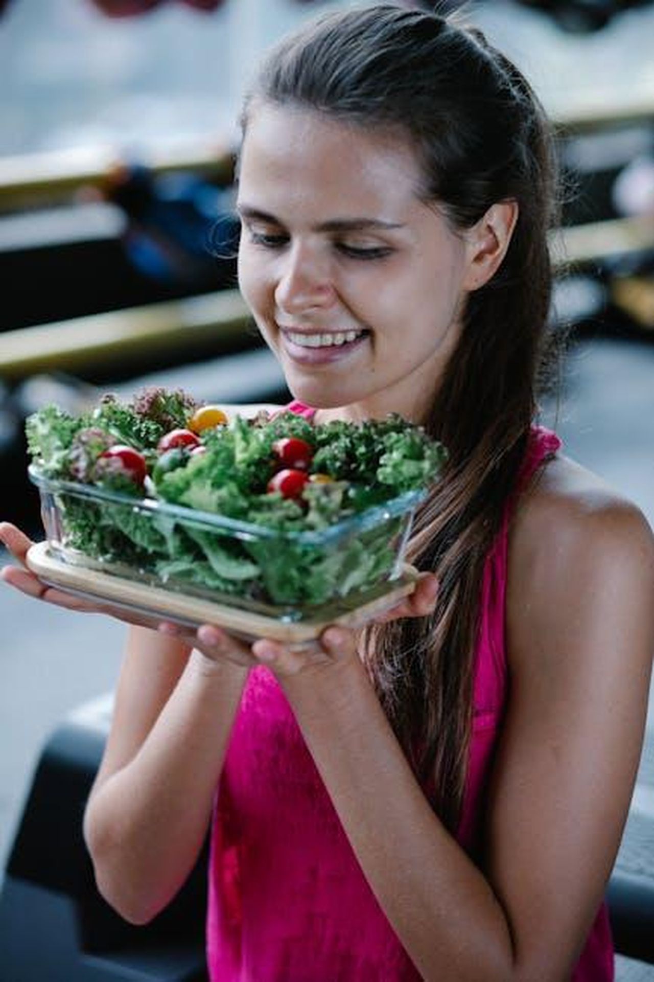 Source: Annushka Ahuja - pexels - Smiling woman in a pink top holding a bowl of fresh salad with cherry tomatoes and greens indoors.