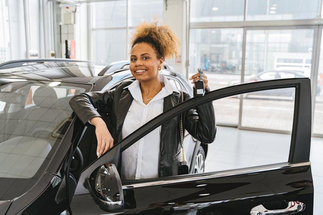Source: Antoni Shkraba - pexels - Smiling woman holding car key in dealership showroom next to new vehicle.