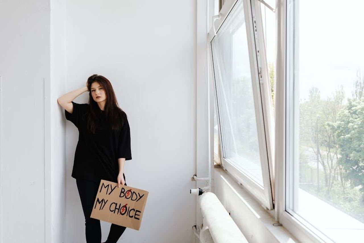 Source: Photo By: Kaboompics.com - pexels - Woman in black holding 'My Body My Choice' sign indoors by a window, highlighting activism.