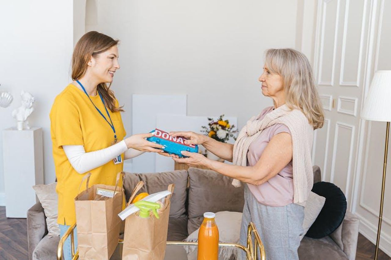 Source: Antoni Shkraba - pexels - Senior woman giving donations to a smiling volunteer inside a cozy living room.