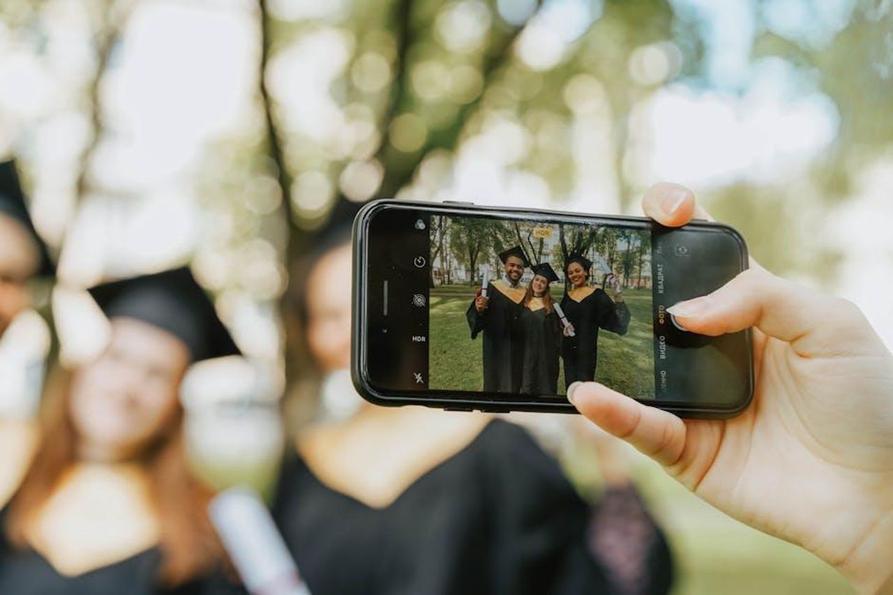 Source: olia danilevich - pexels - A group of graduates taking a photo with a smartphone in a park setting.