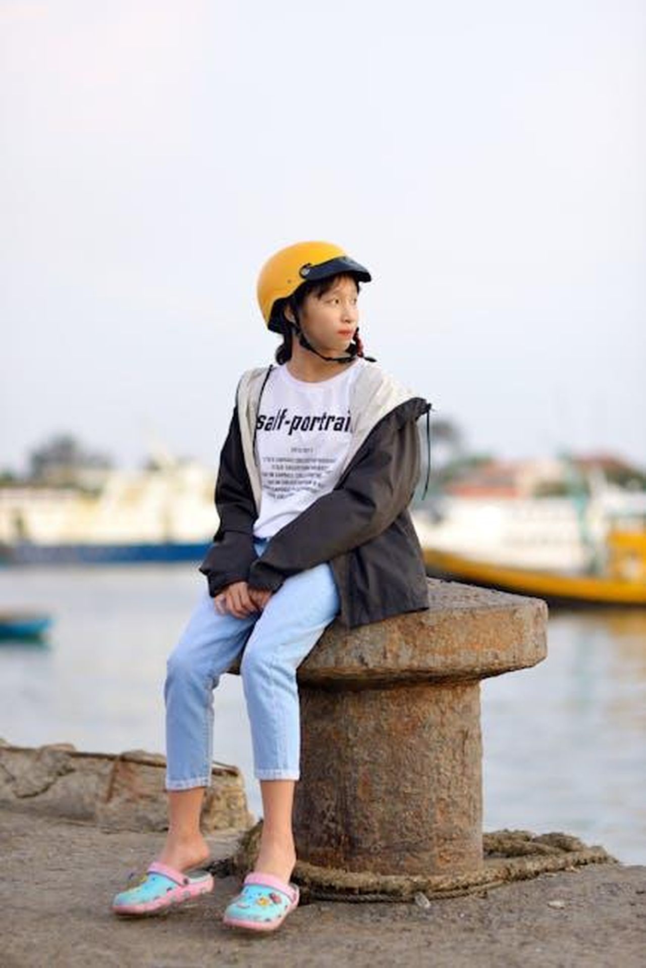 Source: Viễn Đông - pexels - A young girl wearing a helmet sits by the waterfront, looking thoughtful.