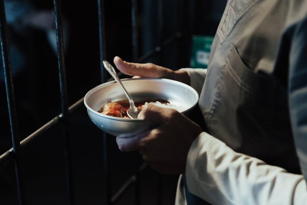 Source: Ron Lach - pexels - Person holding a meal bowl in front of prison bars, suggesting confinement.