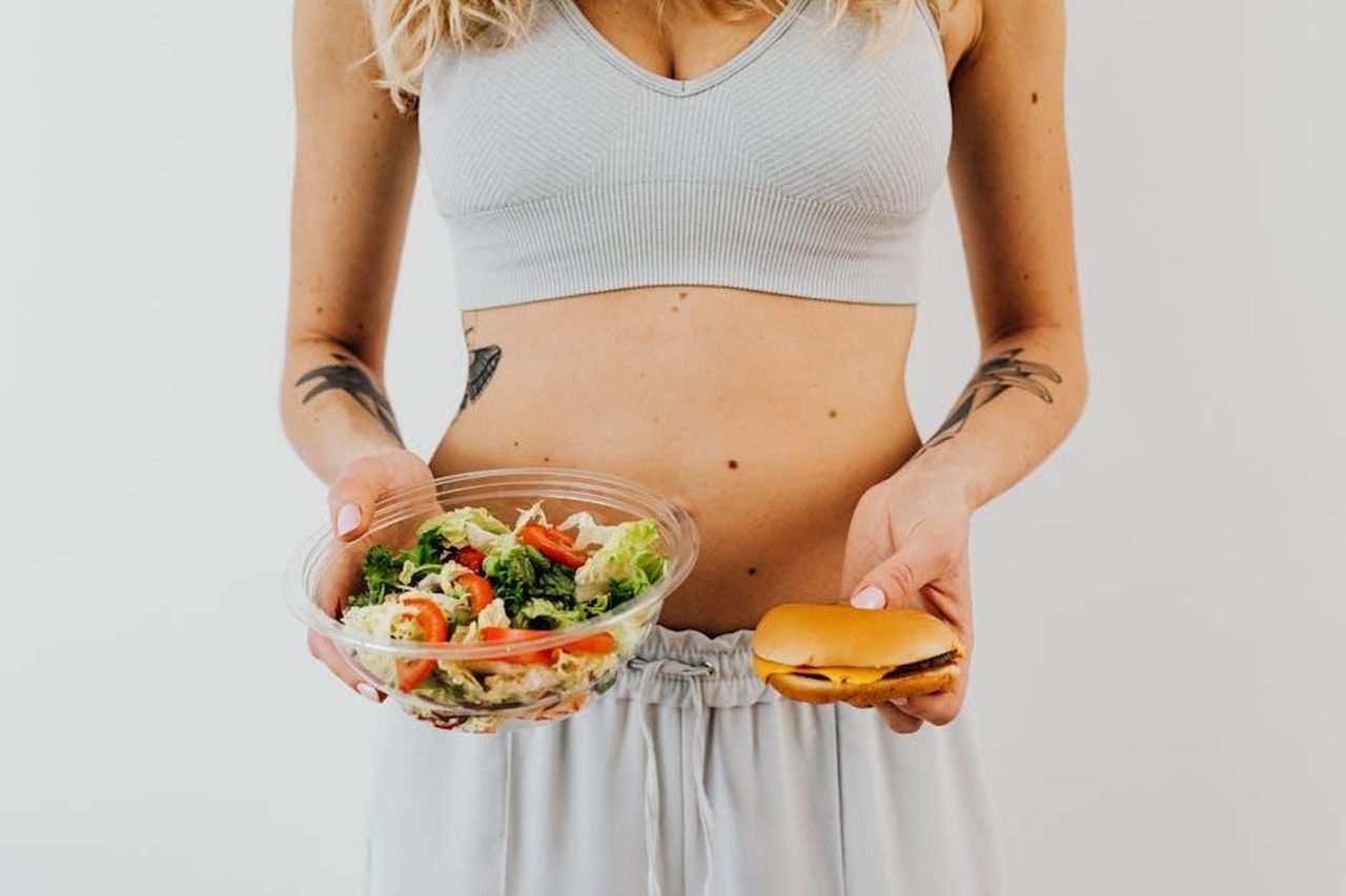 Source: Photo By: Kaboompics.com - pexels - A woman in a sports bra holding a salad and cheeseburger, representing healthy vs junk food choices.