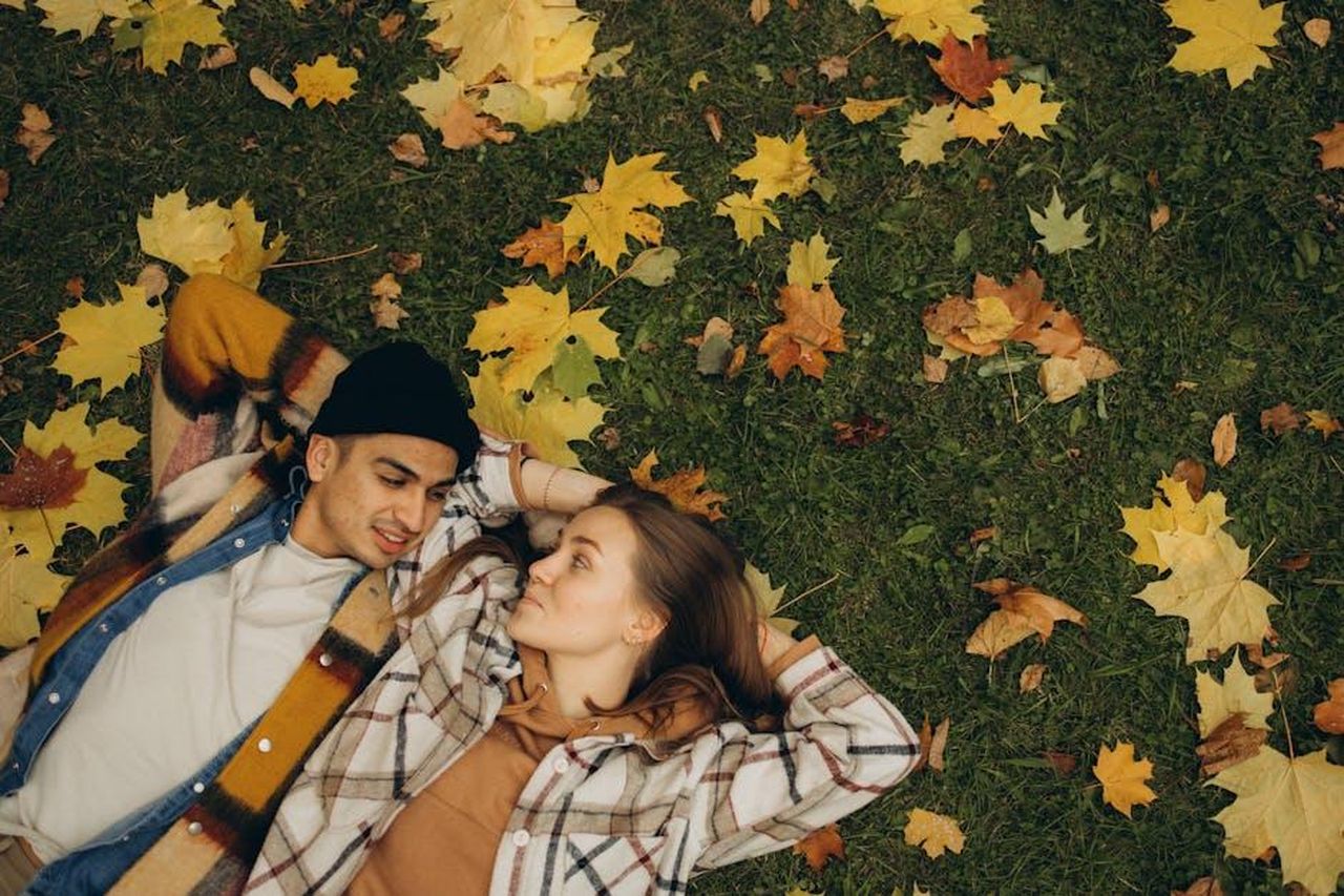 Source: Maksim Goncharenok - pexels - Young couple lying on grass surrounded by autumn maple leaves, embracing seasonal warmth.