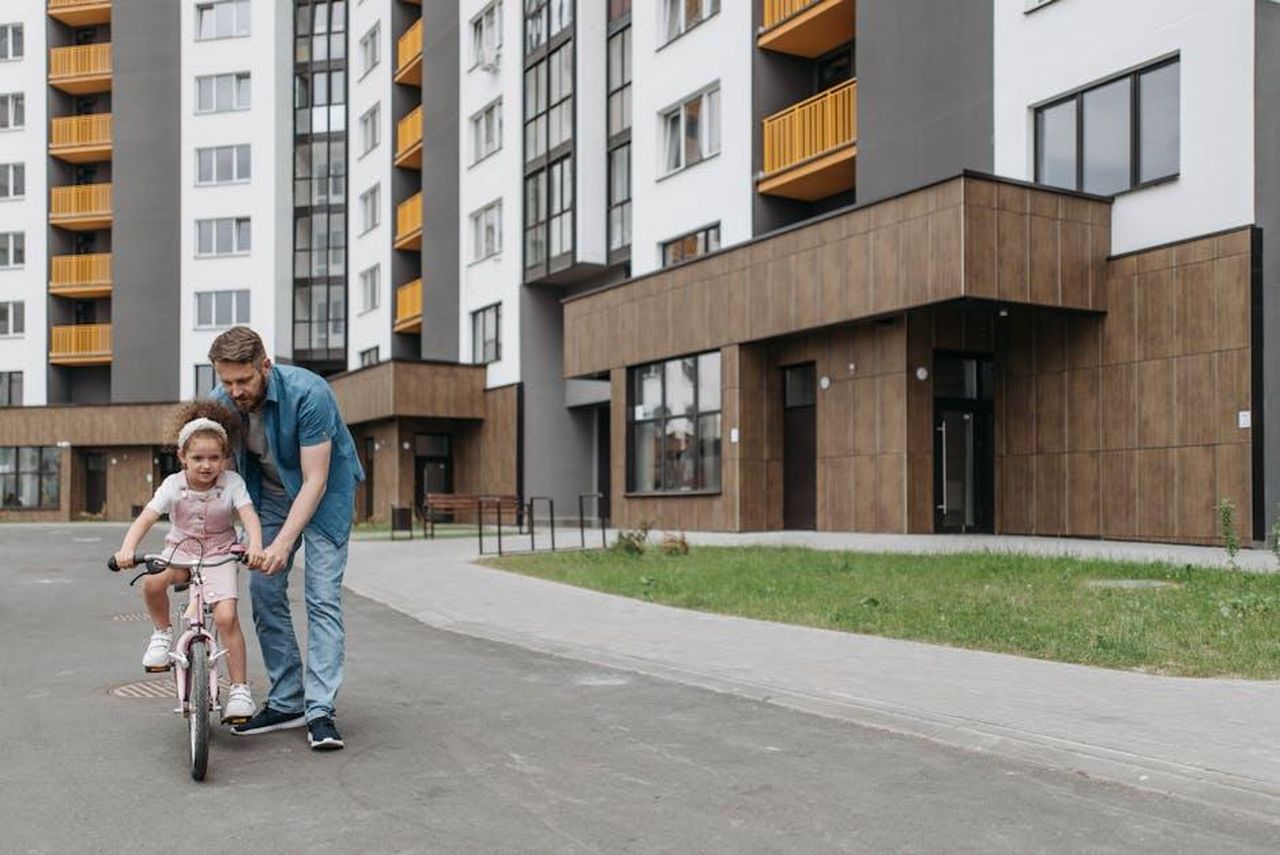Source: Pavel Danilyuk - pexels - Father guiding daughter on a bicycle in front of modern condominiums, symbolizing family bonding and outdoor fun.