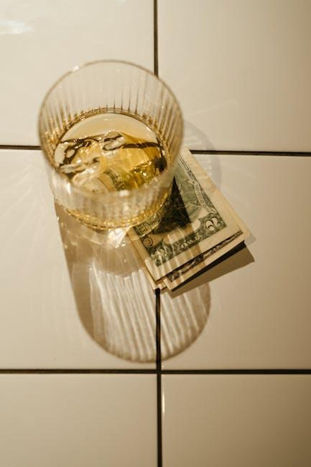 Source: cottonbro studio - pexels - A close-up of a glass of whiskey with ice on top of dollar bills, casting shadows on white tiles.