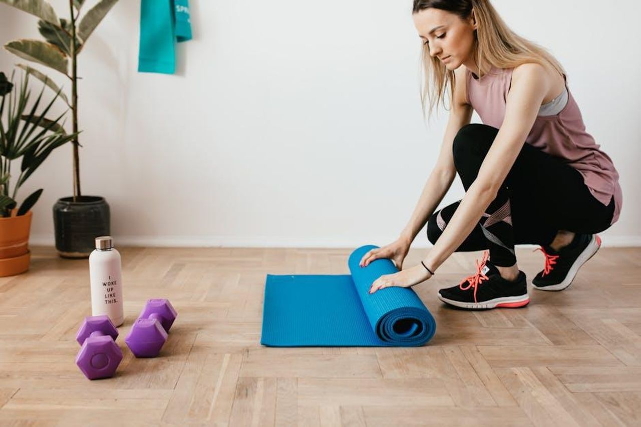 Source: Photo By: Kaboompics.com - pexels - Young woman unfolding yoga mat in a home gym setting with accessories and plants.