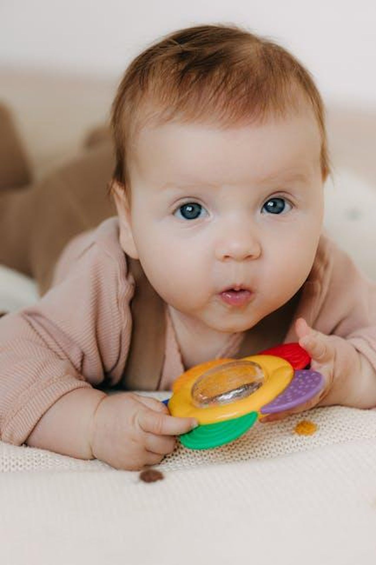 Source: Polesie Toys - pexels - Cute baby holding a colorful sensory toy while lying on a soft surface indoors.