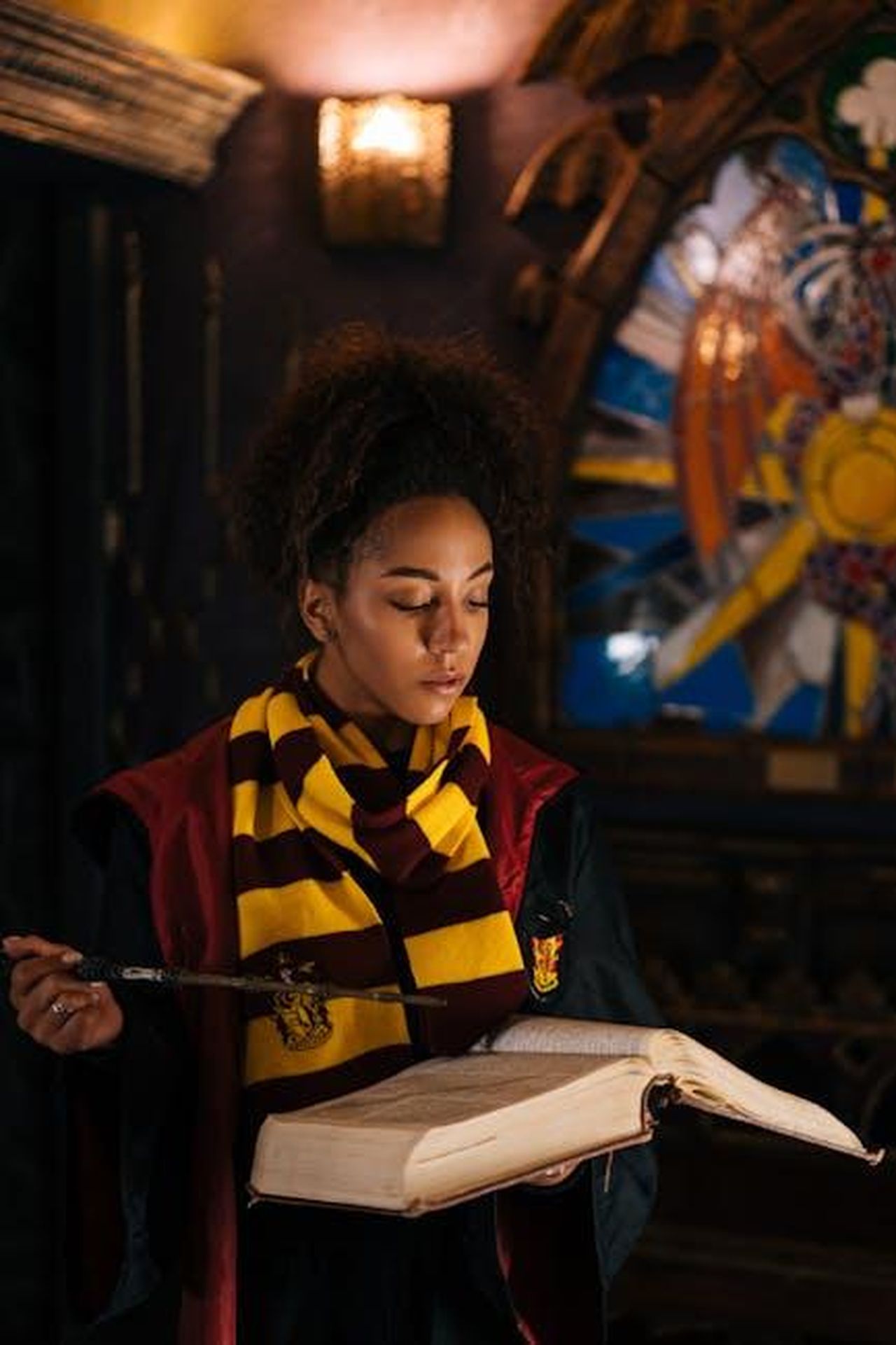 Source: Mikhail Nilov - pexels - African American woman dressed as a wizard reading an open book, holding a wand in a dim, mystical setting.
