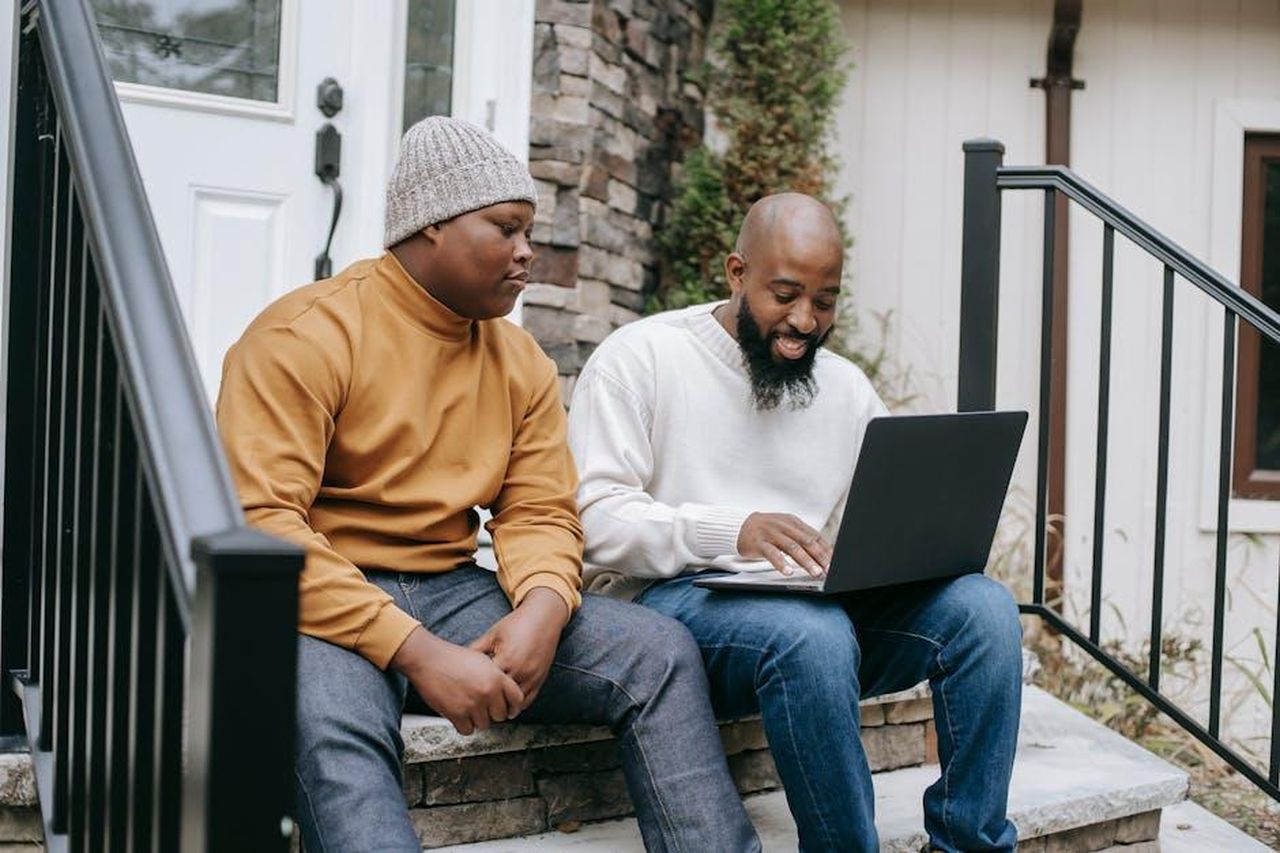 Source: Any Lane - pexels - Positive bearded African American male remote worker sitting on stairs and typing on netbook keyboard with teen son