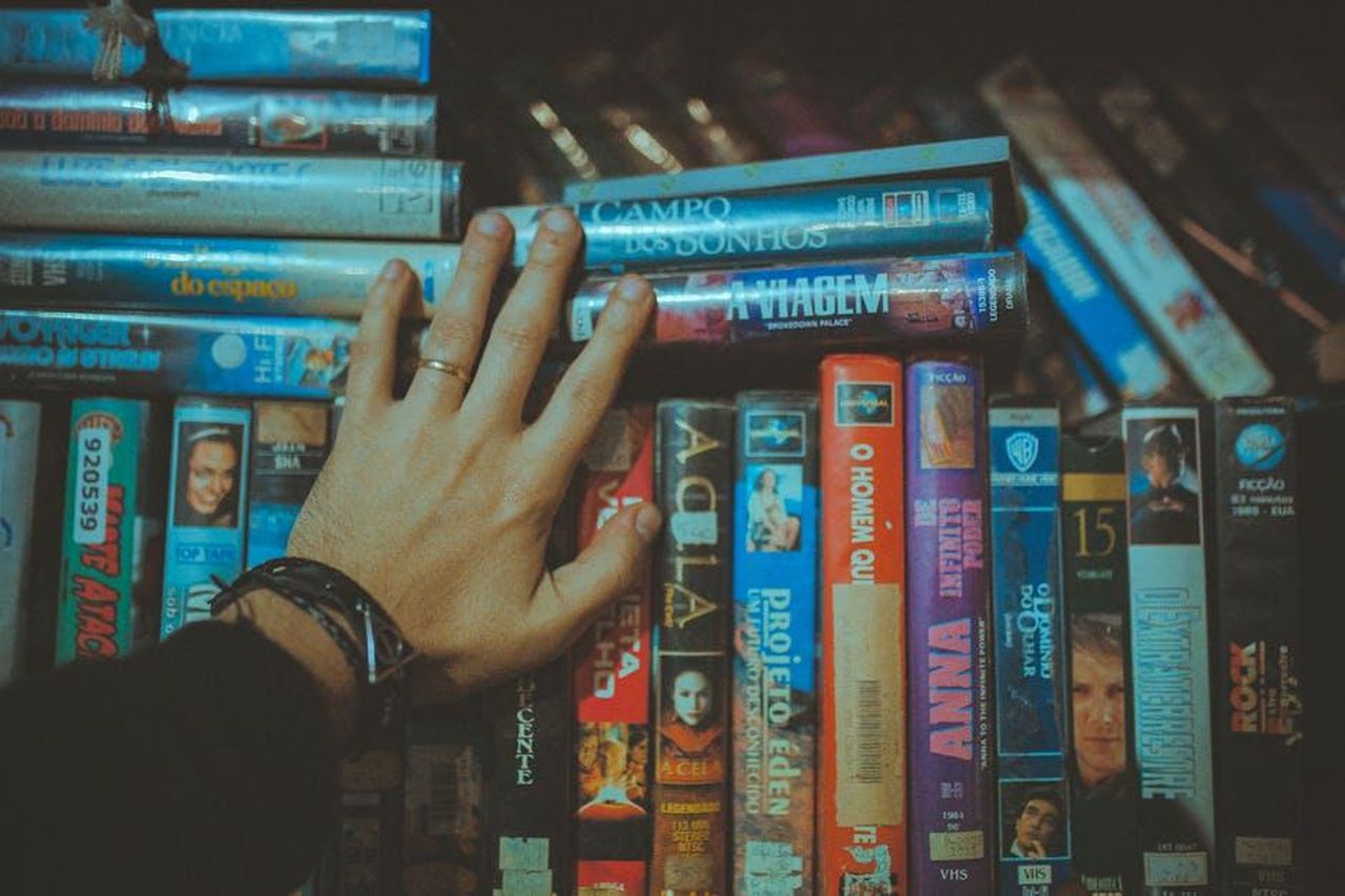 Source: Lucas Pezeta - pexels - A hand reaching for a classic VHS tape from a colorful vintage collection indoors.