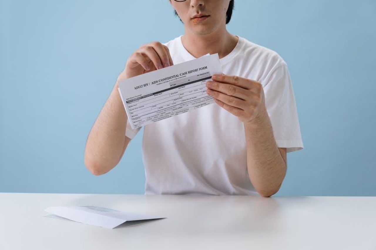 Source: cottonbro studio - pexels - Individual reviewing a confidential HIV/AIDS case report form indoors against a blue background.