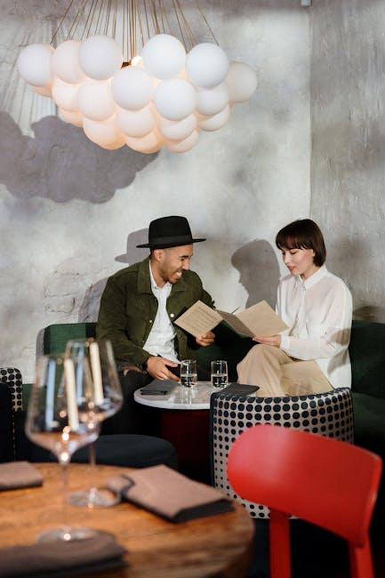 Source: cottonbro studio - pexels - A couple sitting at a table in a stylish café choosing from the menu under a unique ceiling lamp.