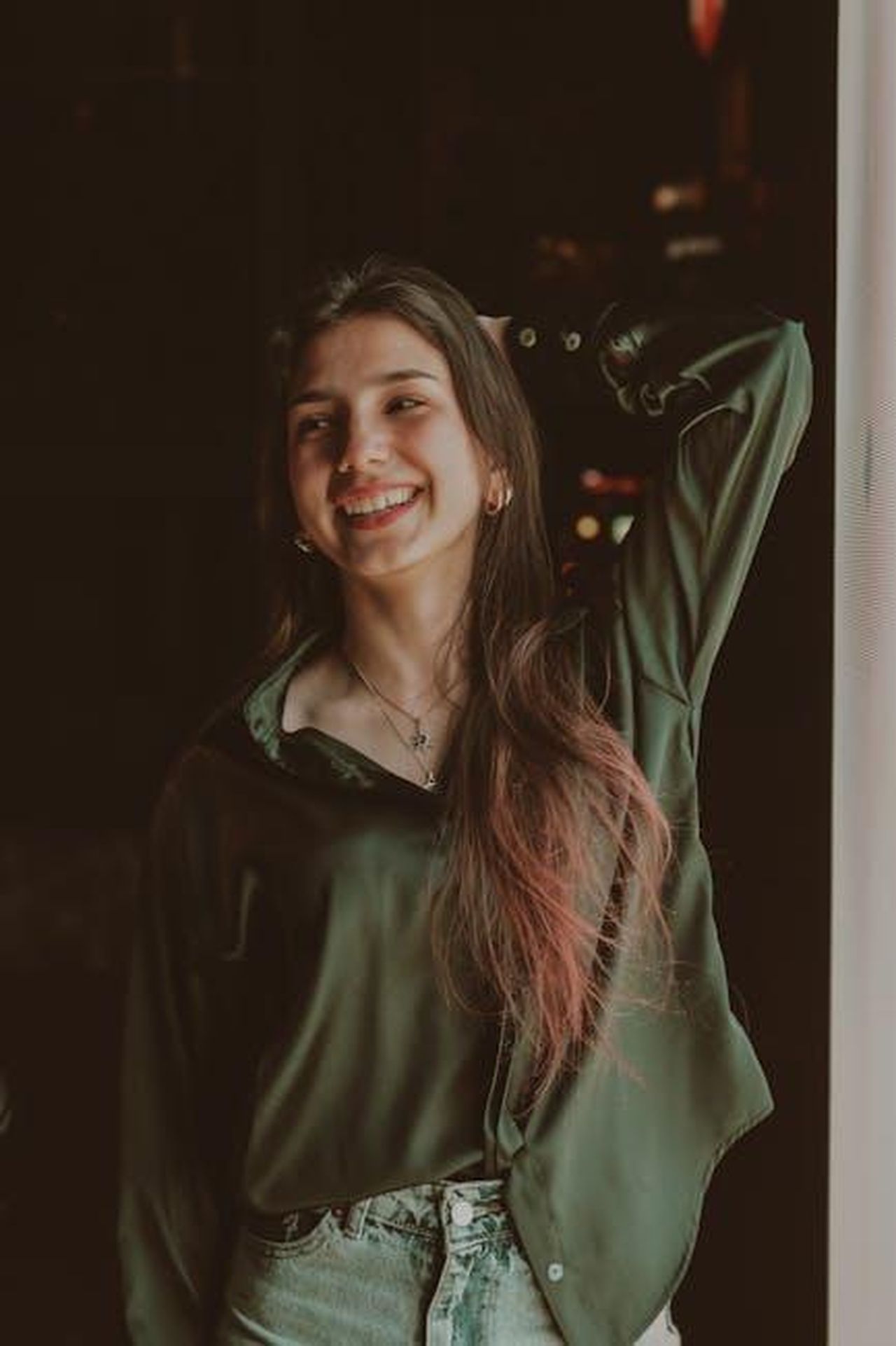 Source: Murat IŞIK - pexels - A joyful woman wearing a green blouse, smiling and posing indoors near a window.