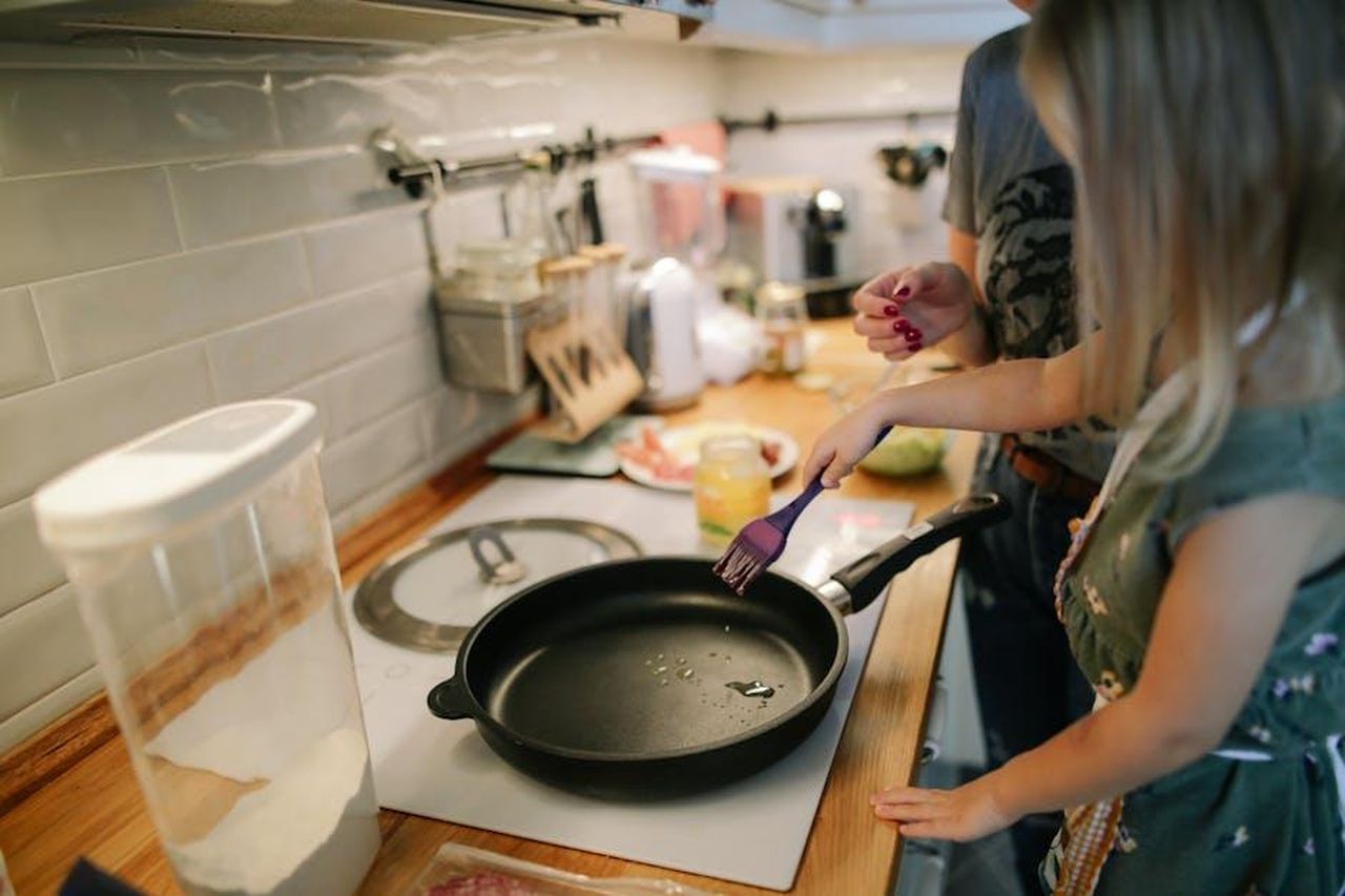 Source: Daria Obymaha - pexels - Young girl cooking with a frying pan under adult supervision in a modern kitchen.