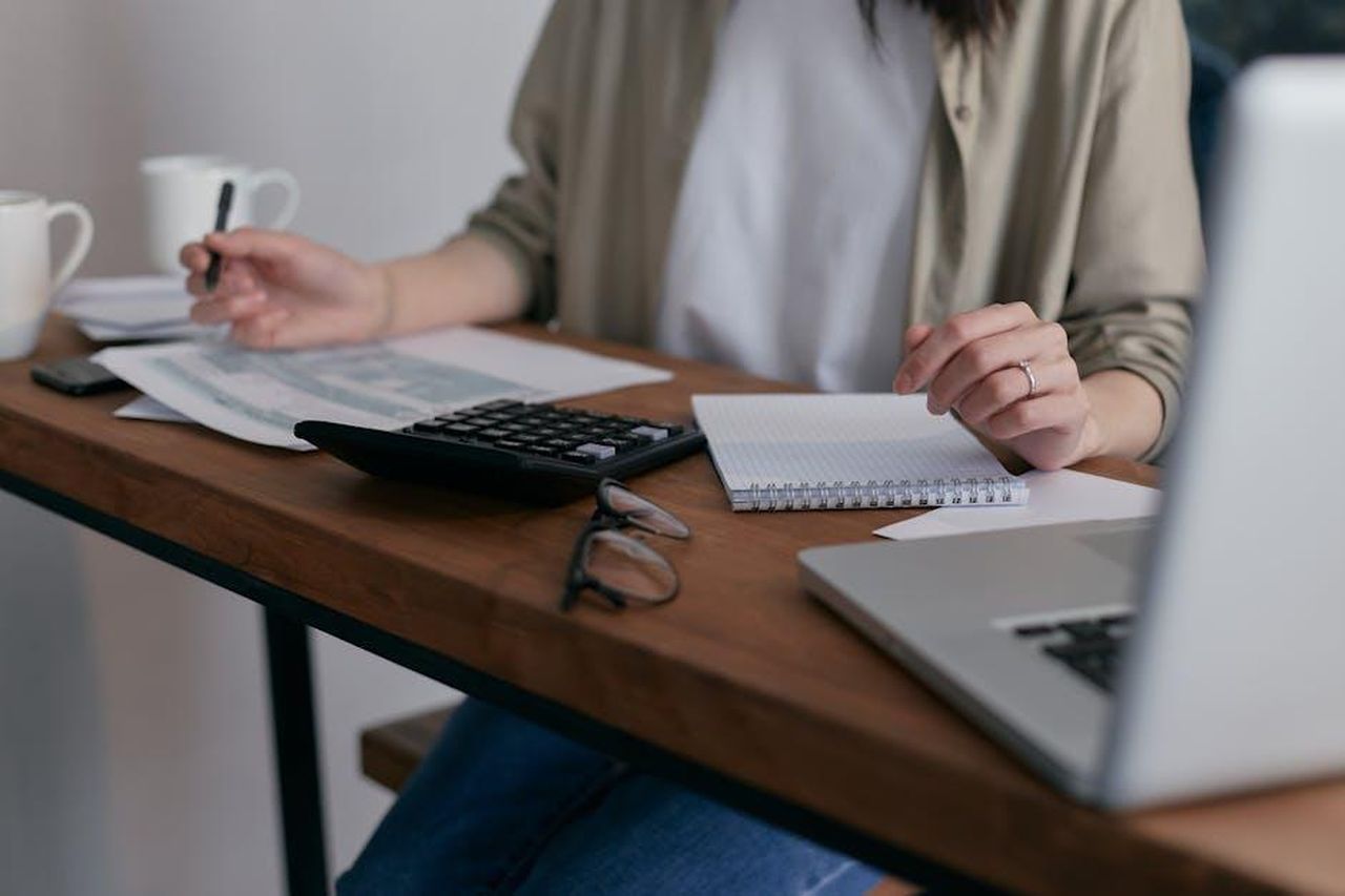 Source: Mikhail Nilov - pexels - A woman manages finances at home, using a laptop and calculator on a wooden desk.