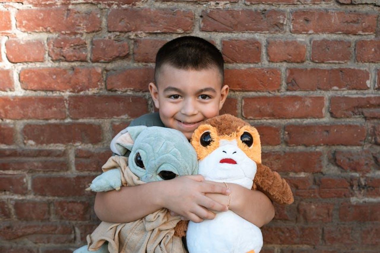 Source: RDNE Stock project - pexels - A cheerful child hugs Grogu and Porg plush toys against a rustic brick wall background.