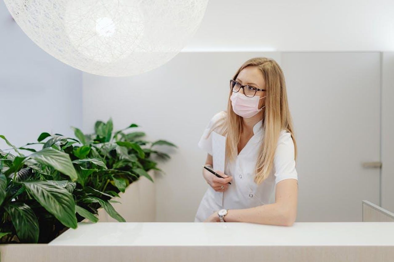 Source: Photo By: Kaboompics.com - pexels - A female healthcare worker in a clinic wearing a face mask, standing at the reception desk.