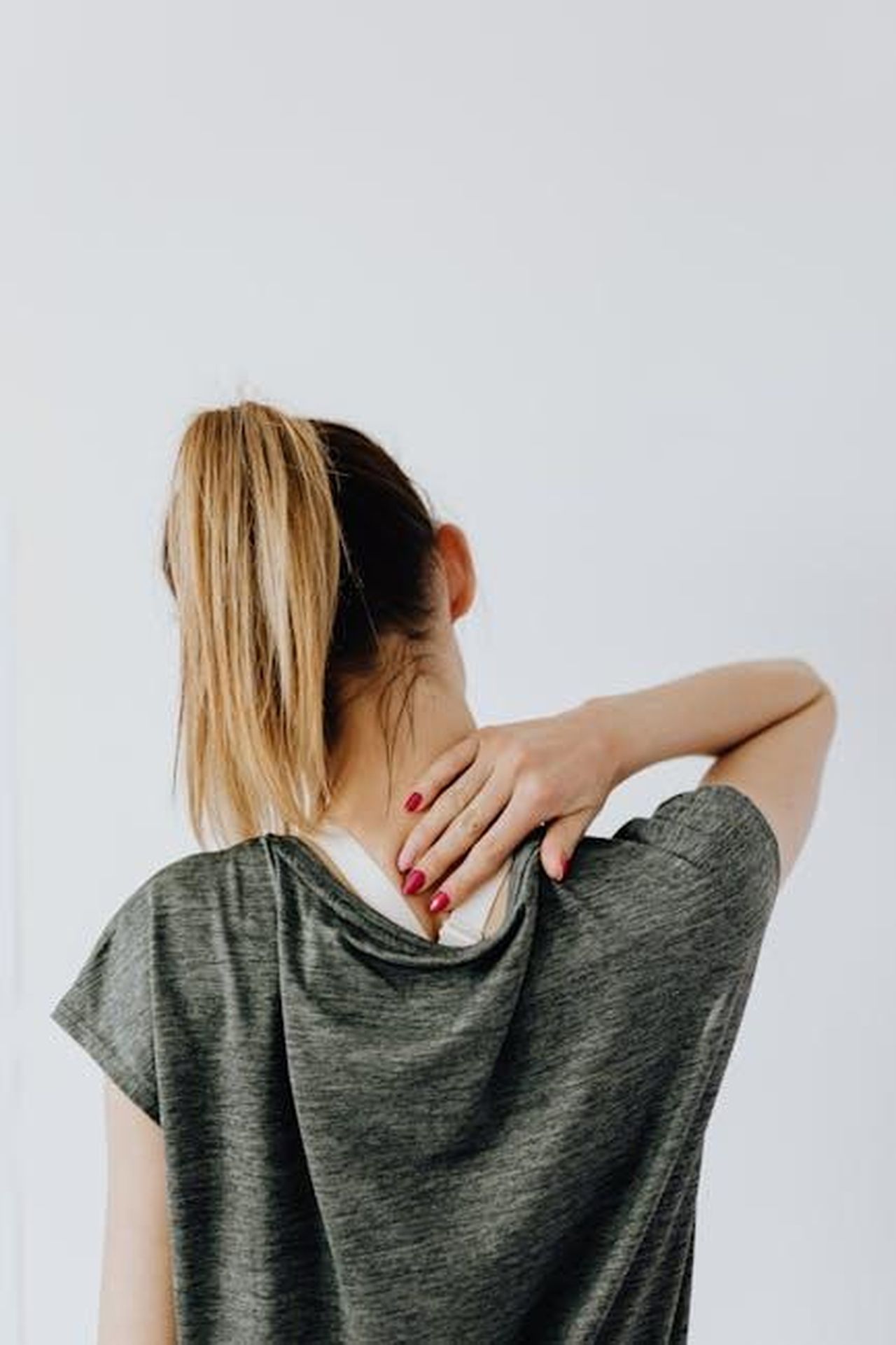 Source: Photo By: Kaboompics.com - pexels - Back view of blond faceless lady in casual t shirt and manicure demonstrating pain spot on back while sitting near white wall