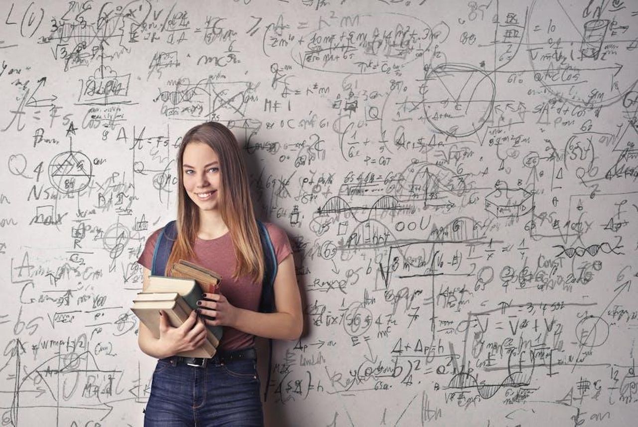 Source: Andrea Piacquadio - pexels - Young woman holding books standing in front of a chalkboard filled with mathematical formulas.