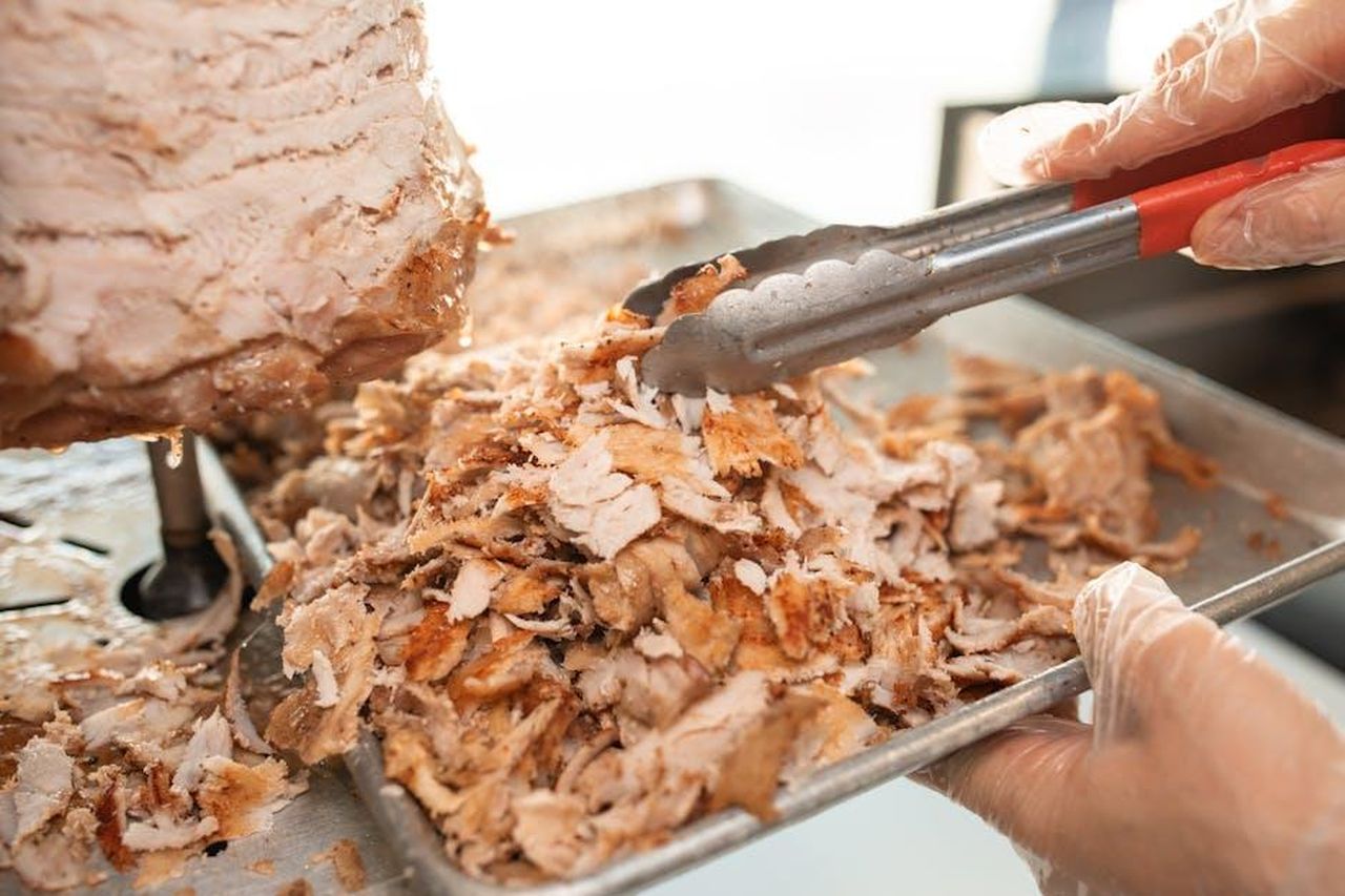 Source: RDNE Stock project - pexels - Freshly sliced shawarma meat on a tray being prepared with tongs in a kitchen setting.
