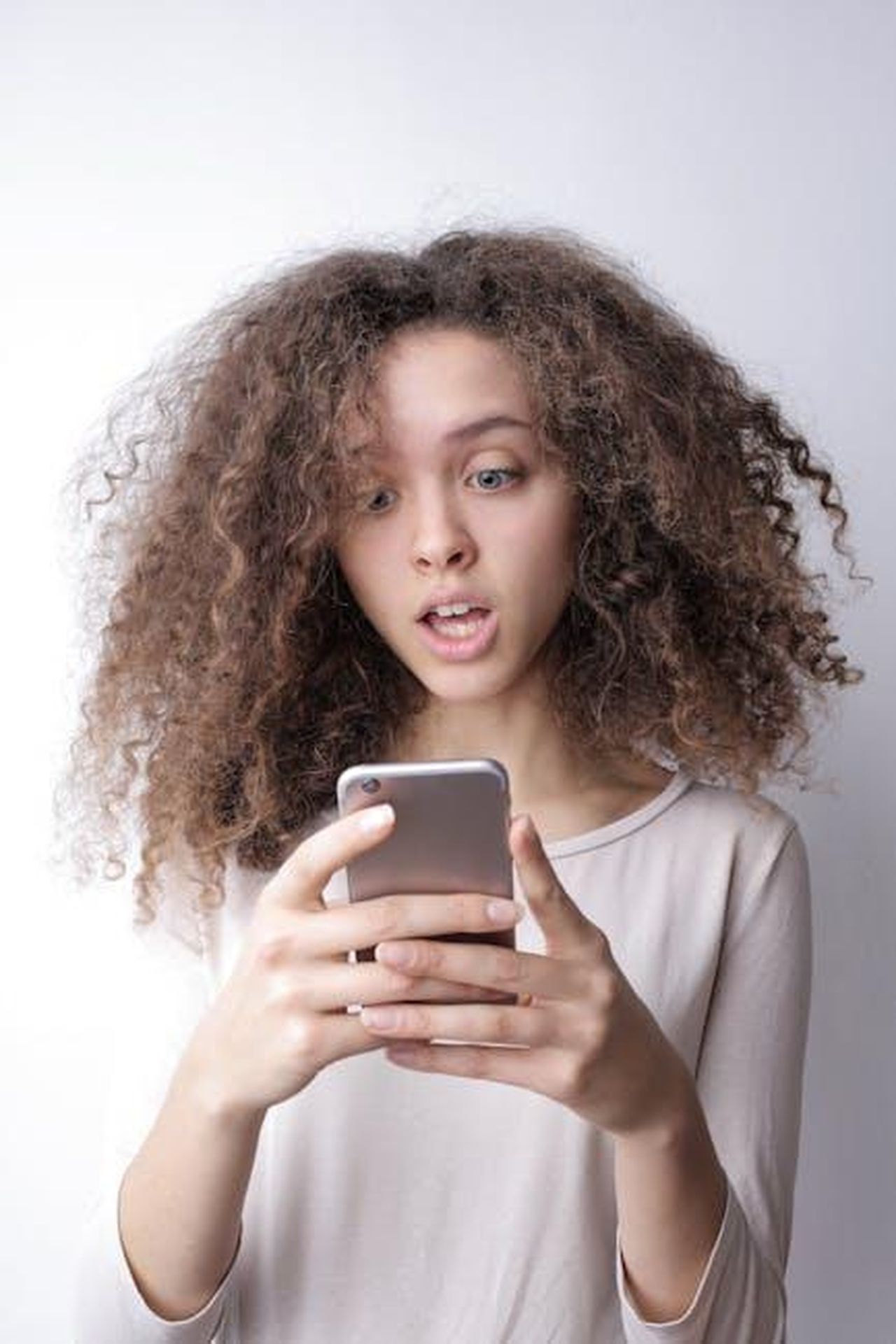 Source: Andrea Piacquadio - pexels - A young woman with curly hair looks surprised while reading something on her smartphone indoors.