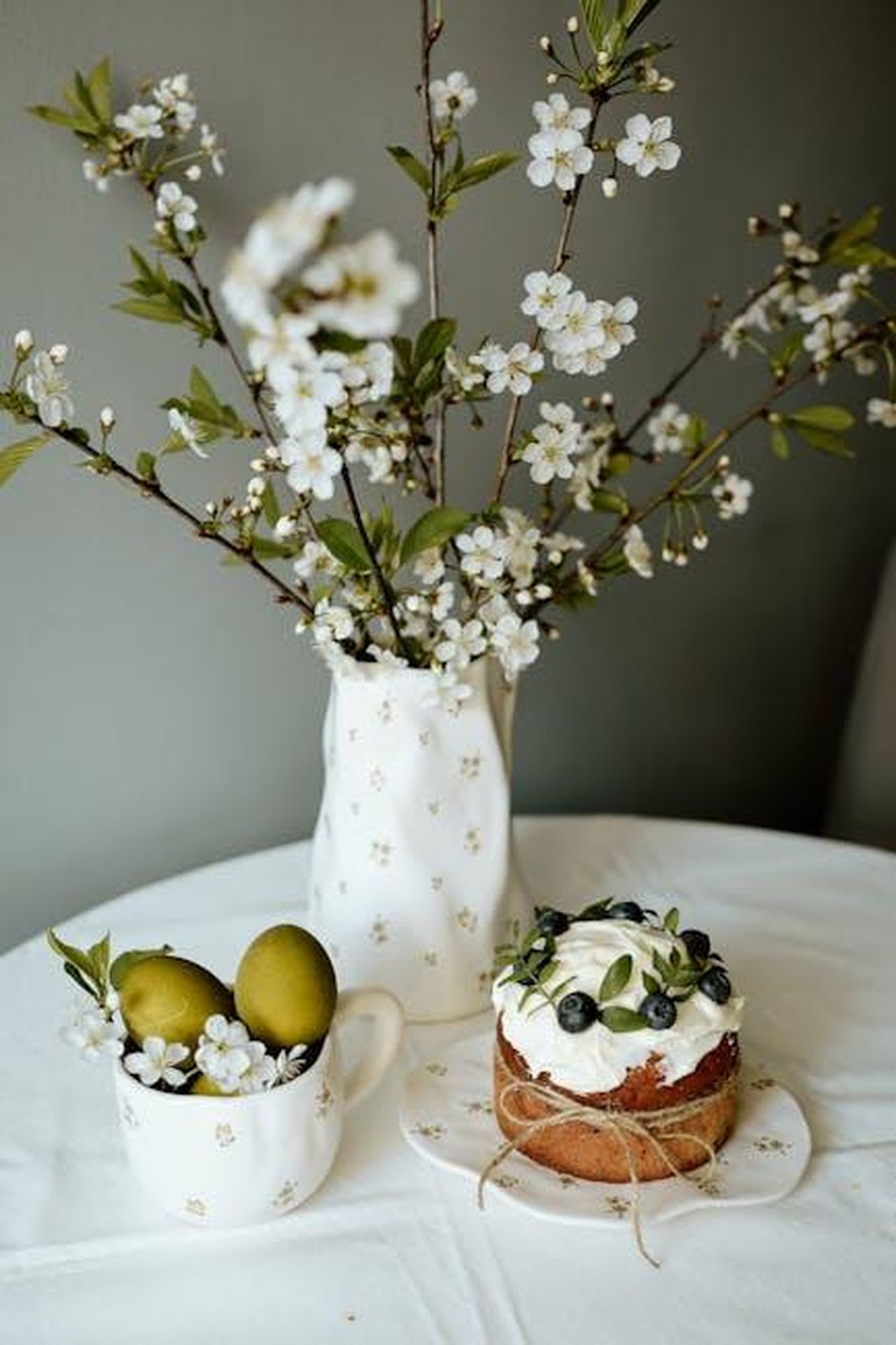 Source: Liana Tril' - pexels - Elegant table setting with blooming flowers, fruit-filled cup, and decorated cake.