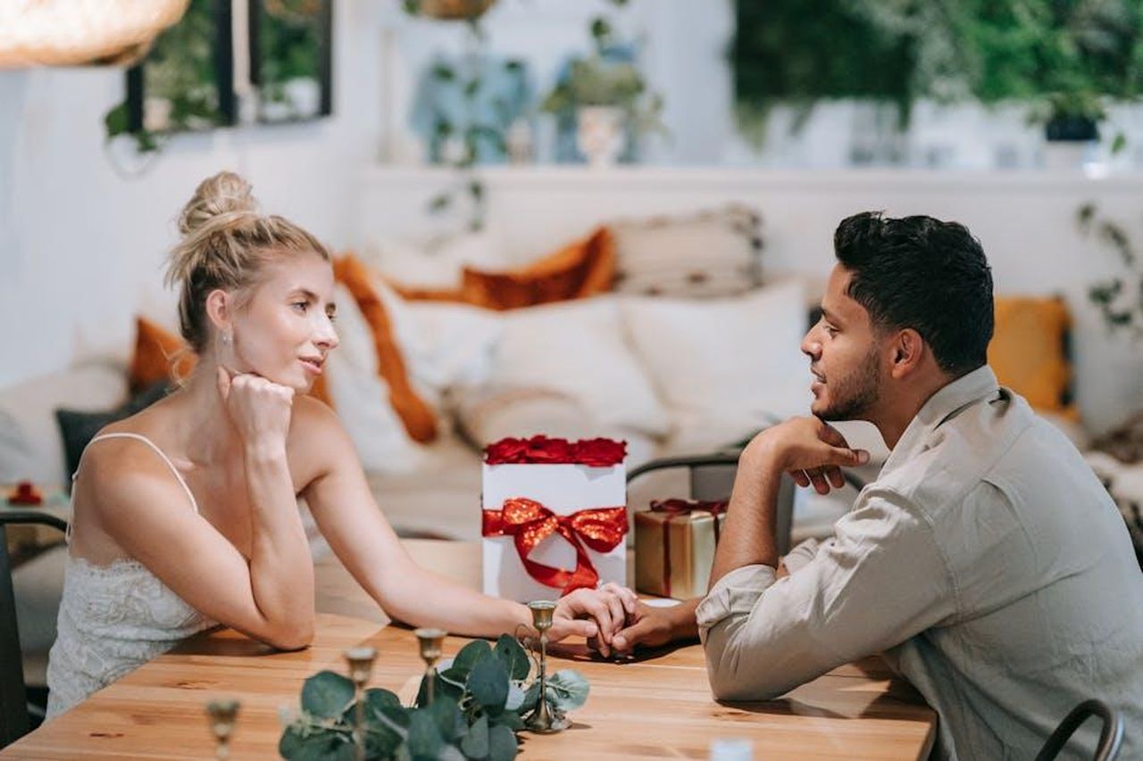 Source: Anna Pou - pexels - A loving couple enjoying a romantic date, holding hands indoors with a gift on the table.